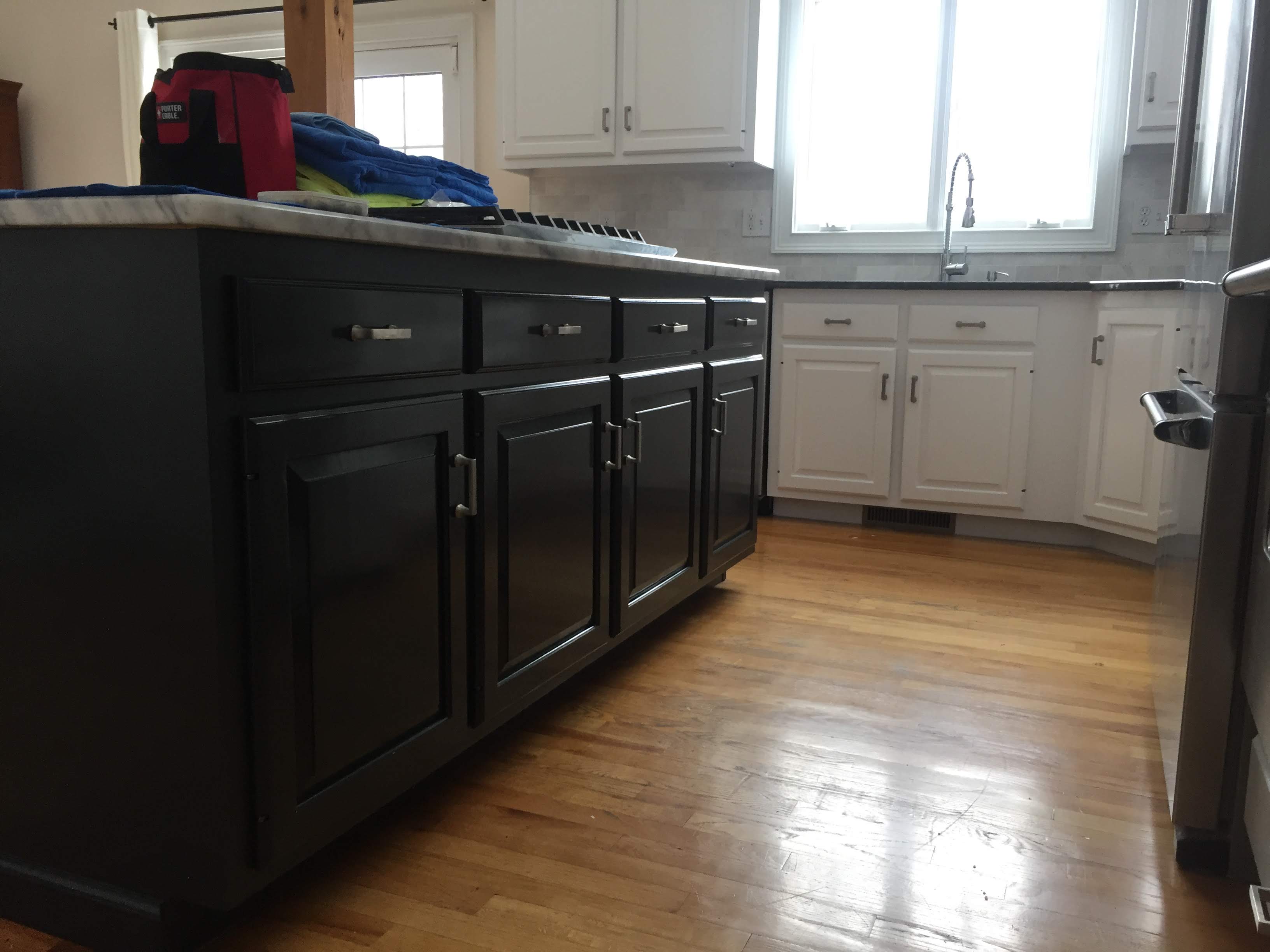 Kitchen island with dark cabinetry and marble countertop next to white cabinets, granite counters, and a farmhouse sink under a window by Rome Leite Home Improvement in Danbury, CT